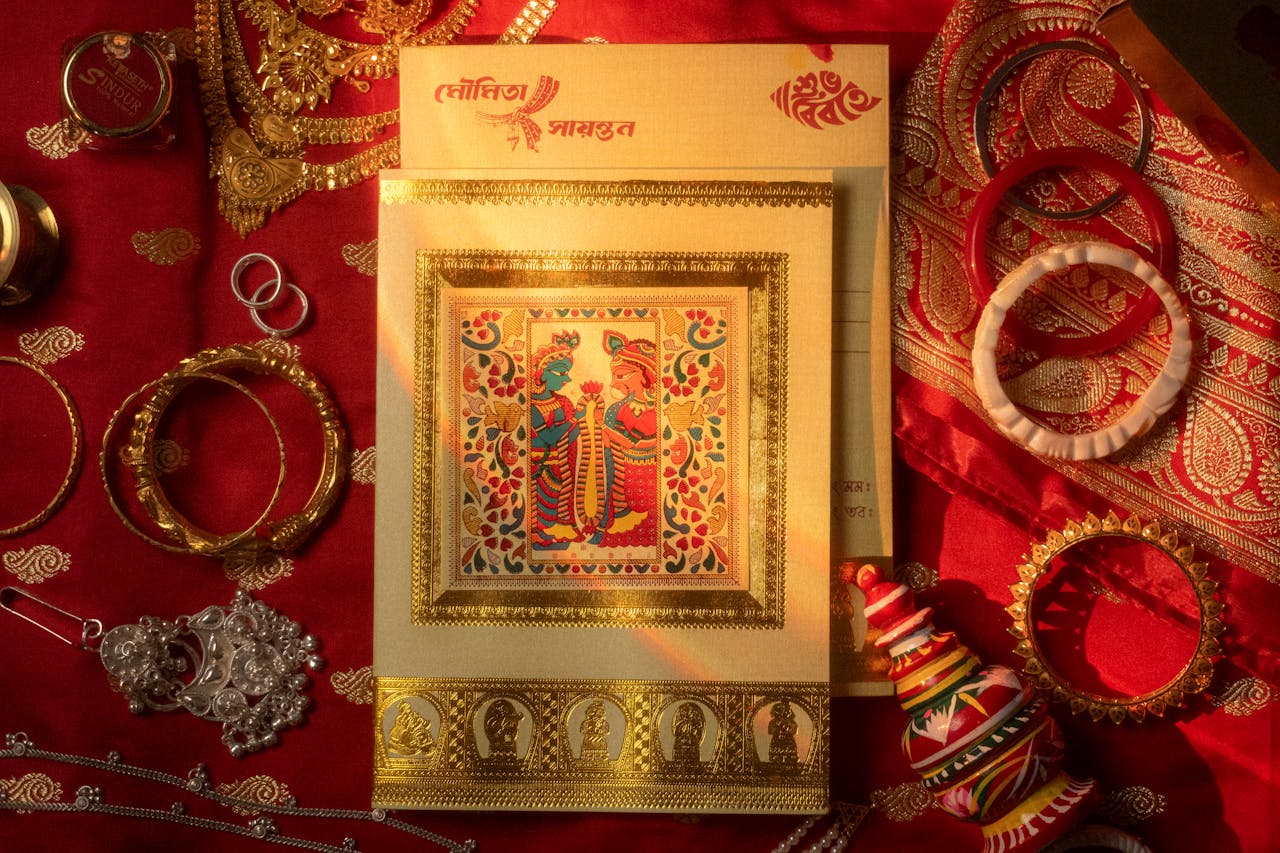A beautiful traditional Indian wedding card with intricate design surrounded by jewelry on a red fabric.